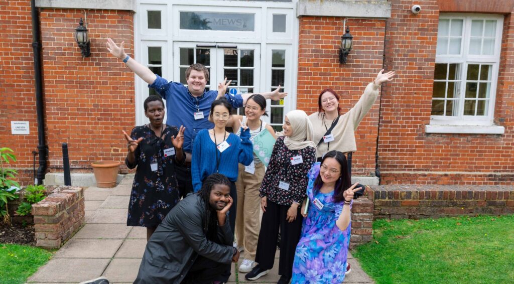 Group of 8 PhD students posing with arms raised on grey path in front of red-bricked building.