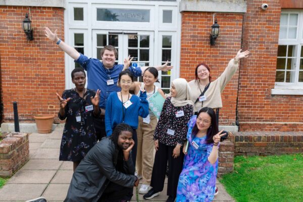 Group of 8 PhD students posing with arms raised on grey path in front of red-bricked building.