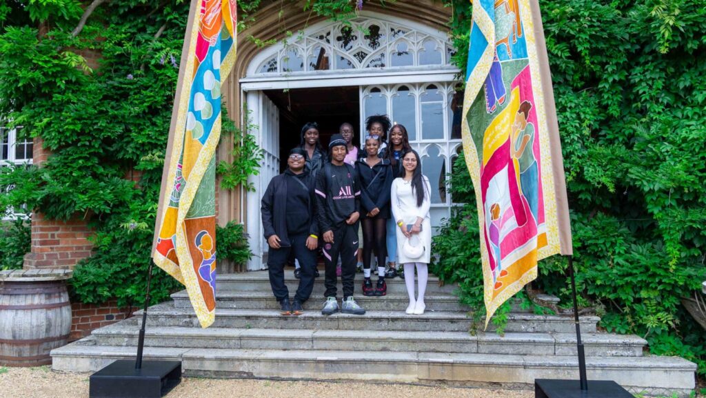 Group of 8 teenagers stand in between two colourful silk flags blowing in the wind, in front of entrance to red brick building.