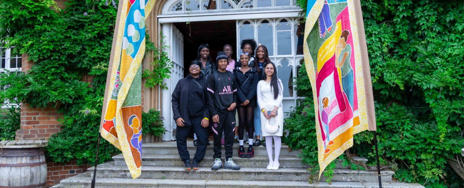 Group of 8 teenagers stand in between two colourful silk flags blowing in the wind, in front of entrance to red brick building.