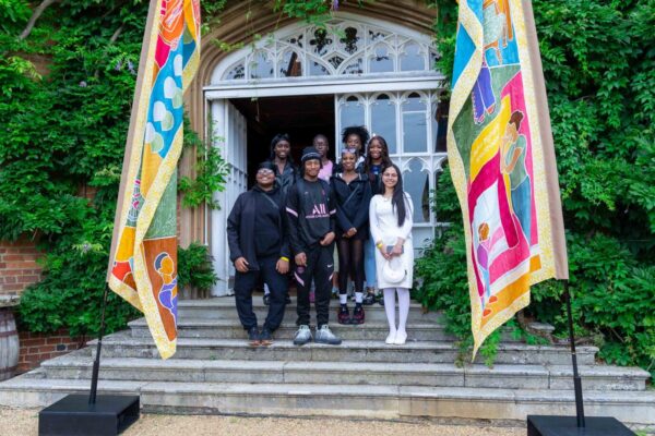 Group of 8 teenagers stand in between two colourful silk flags blowing in the wind, in front of entrance to red brick building.