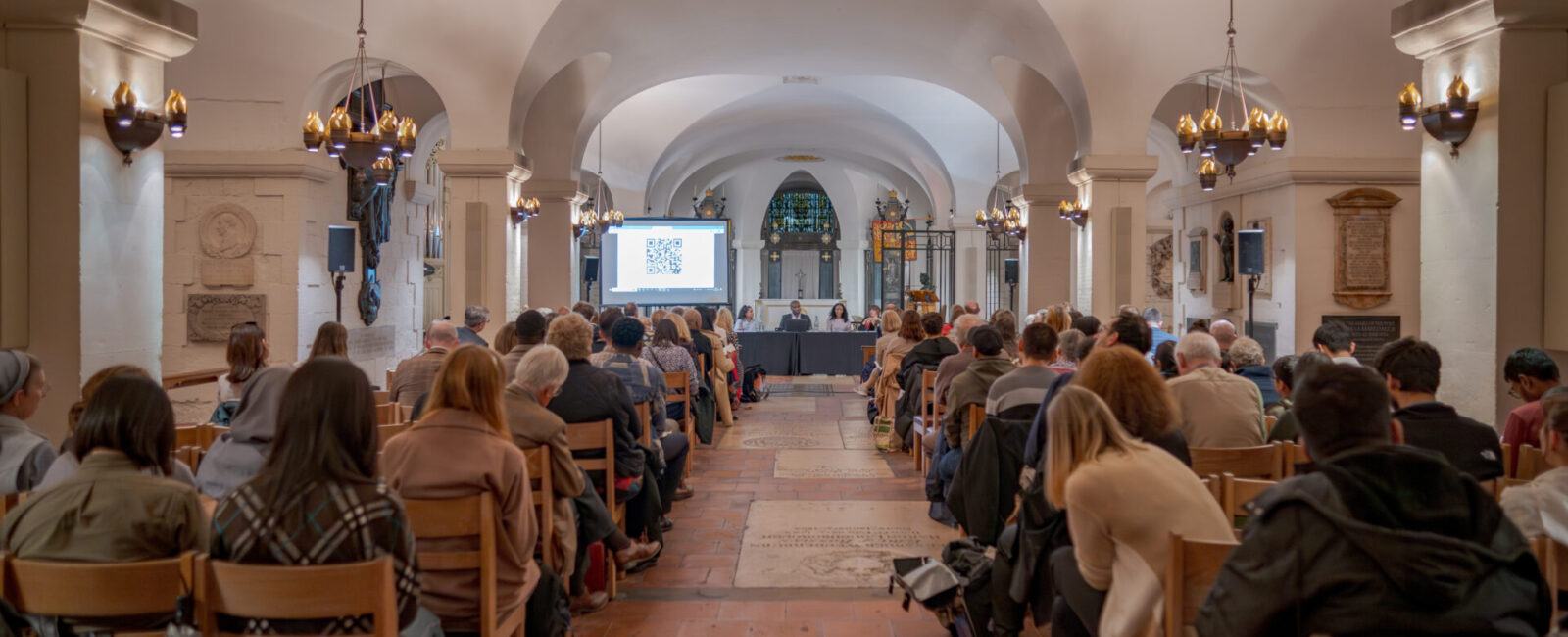 Crowd of people gather in the crypt of St Paul's Cathedral, watching and listening to panel of five members. There is a screen with a QR code in the background, as well as the altar of the crypt in the cathedral.