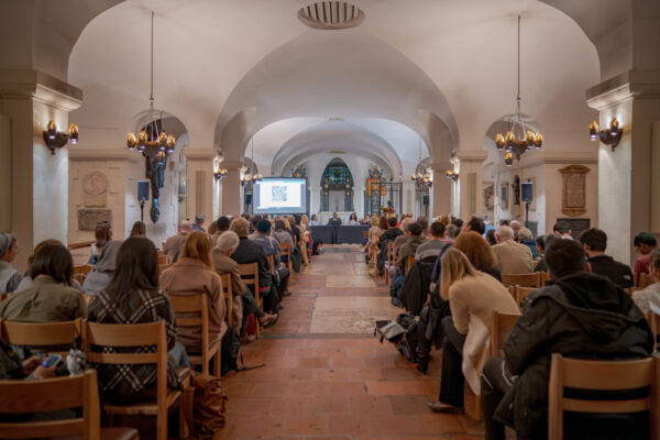 Crowd of people gather in the crypt of St Paul's Cathedral, watching and listening to panel of five members. There is a screen with a QR code in the background, as well as the altar of the crypt in the cathedral.