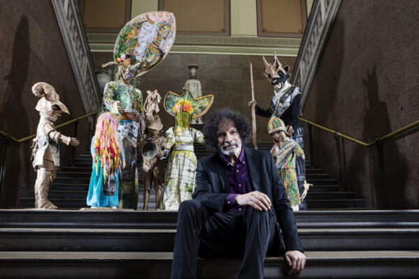 Artist Hew Locke sitting in front of The Watchers, a series of carnivalesque figures installed around the museum galleries.